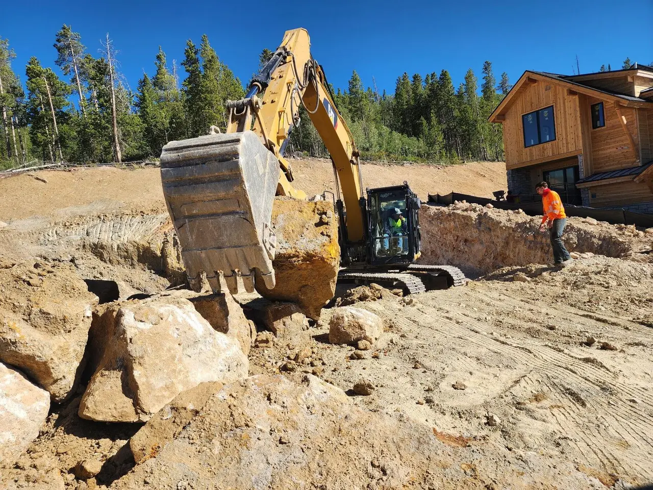 An excavator lifting a boulder out of a dig while a crew member watches