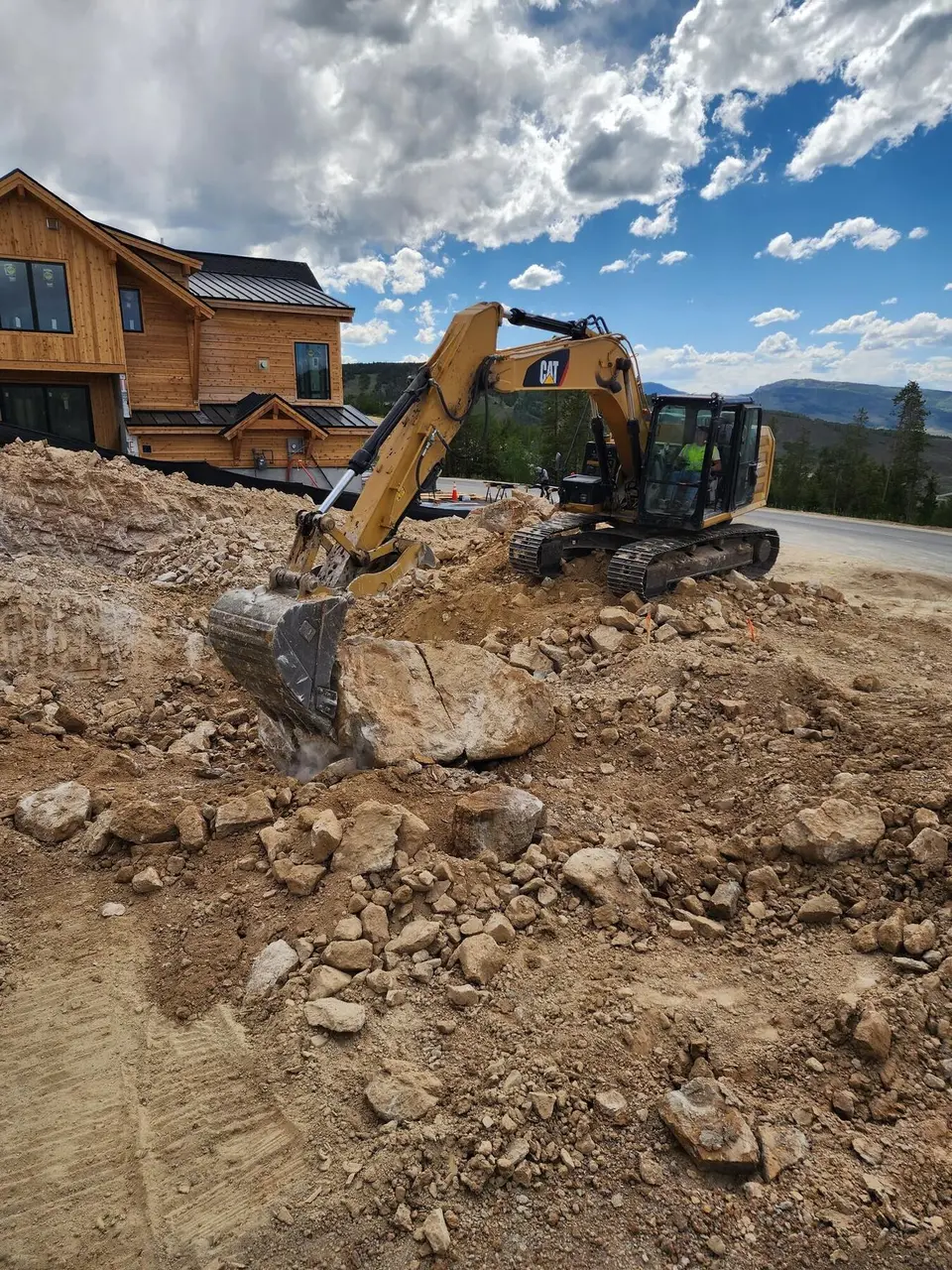 An excavator bucket splitting a large boulder on a cleared lot