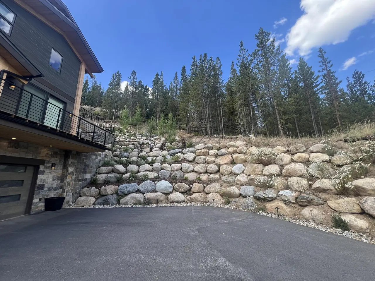 A tiered boulder retaining wall climbing a forested slope beside a driveway