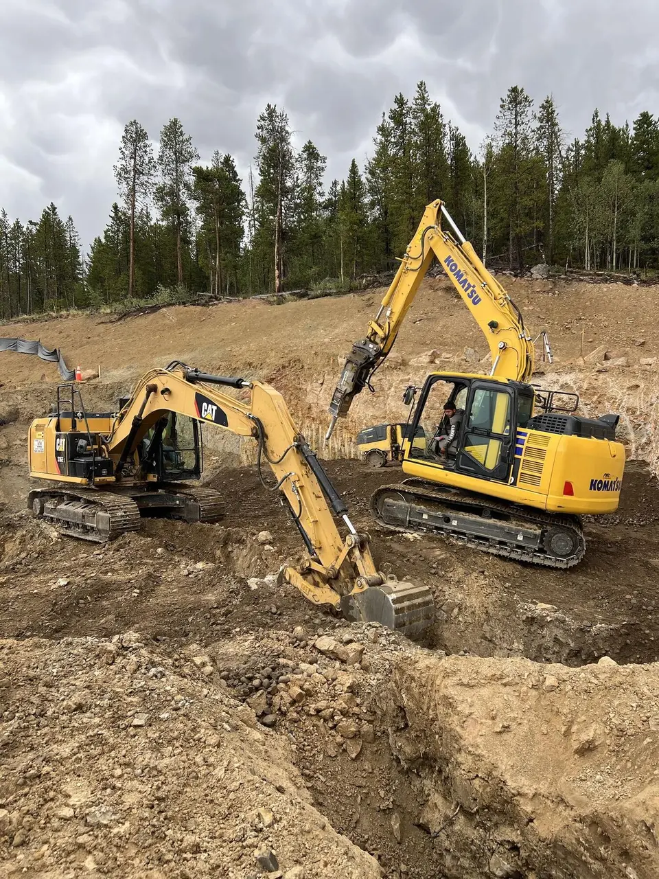 Two excavators at work in a foundation dig, one breaking rock with a hydraulic hammer